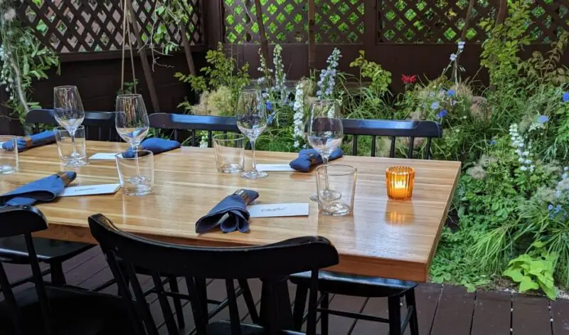 A set table with wine glasses and a candle in a cozy, plant-filled outdoor dining area at Olmsted restaurant