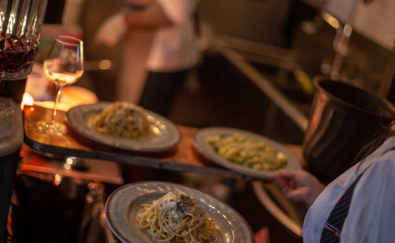 A server carrying plates of pasta in a restaurant kitchen with a glass of wine nearby