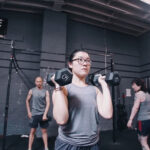 A focused young woman lifting dumbbells in a gym, showcasing determination and strength, while other gym members are seen in the background