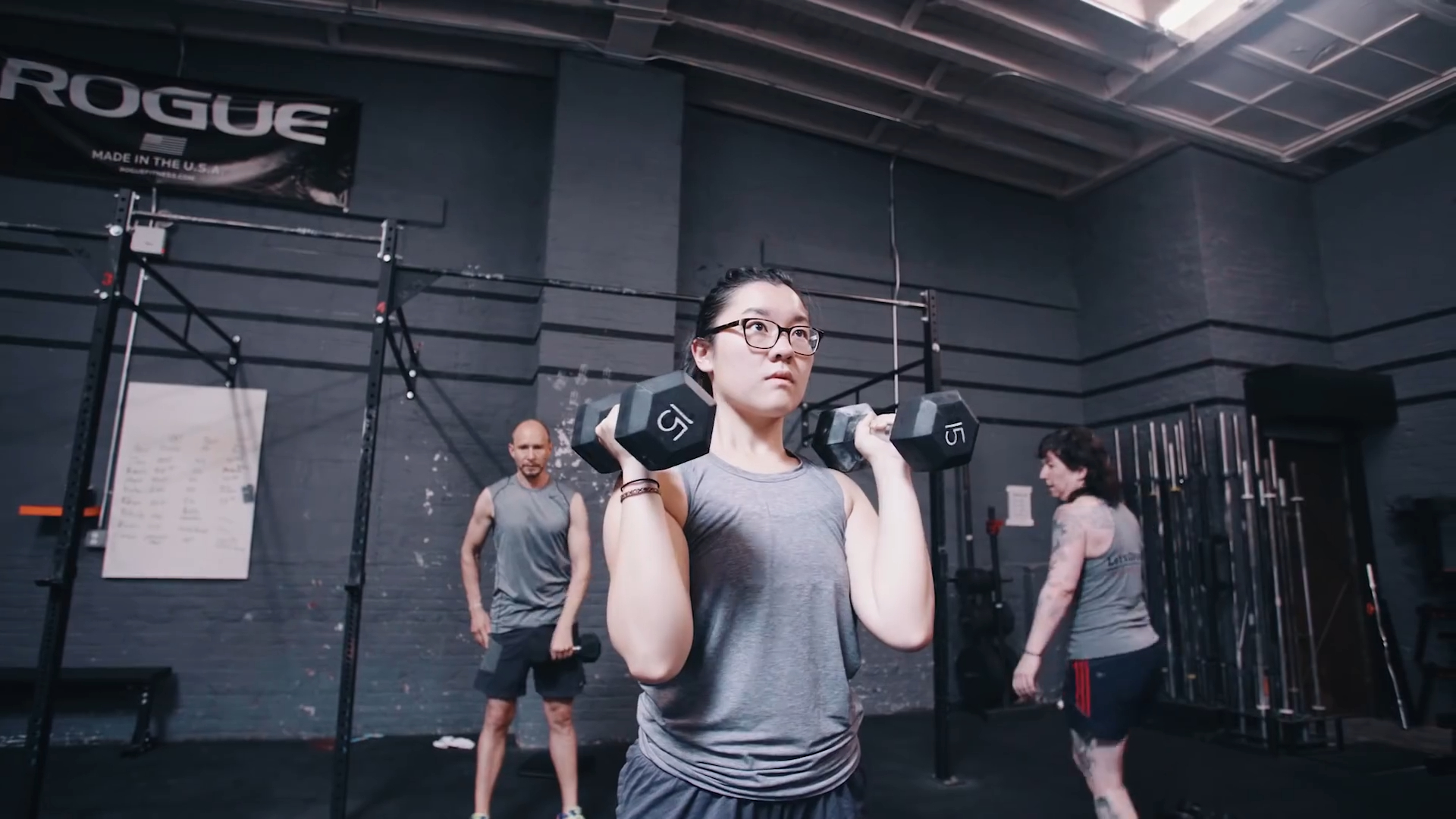 A focused young woman lifting dumbbells in a gym, showcasing determination and strength, while other gym members are seen in the background