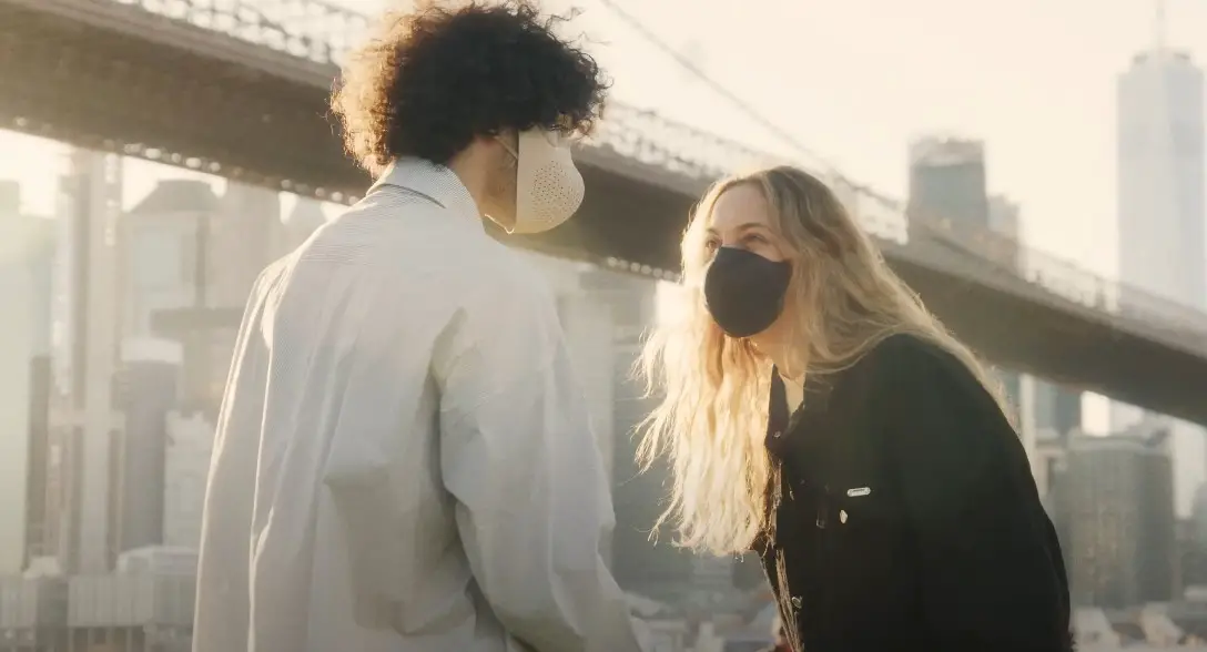 Two individuals converse amidst the urban backdrop of New York City, wearing protective face masks